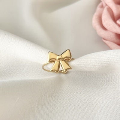 Anel dourado em forma de laço sobre tecido branco com tecido rosa ao fundo