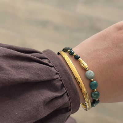 Duas pulseiras no pulso, metal dourado martelado e contas verdes e cinzentas em fundo desfocado e camisa castanha