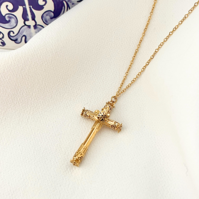 Colar dourado com crucifixo detalhado sobre tecido branco, decoração azul ao fundo