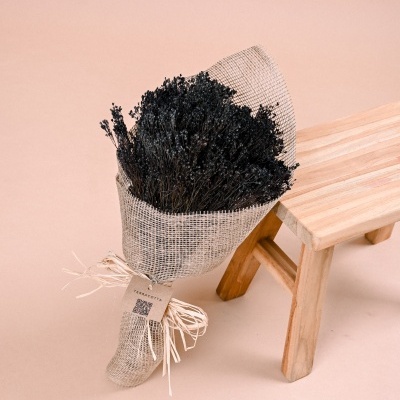 Bouquet de flores secas negras em tecido juta com laço e etiqueta junto a banco pequeno de madeira clara