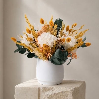 Vaso branco com flores secas amarelas, laranjas, verdes e brancas sobre bloco de pedra clara