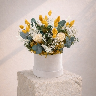 Arranjo floral branco e amarelo em caixa redonda branca sobre bloco de pedra.