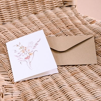 Cartão branco com flores e envelope bege sobre cadeira de vime