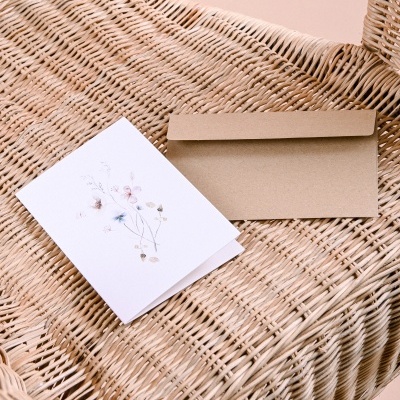 Cartão branco com ilustração floral e envelope castanho sobre cadeira de vime natural