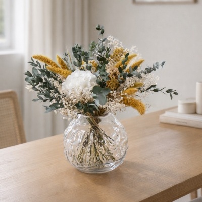 Vaso de vidro com flores secas amarelas, brancas e folhas verdes sobre mesa de madeira