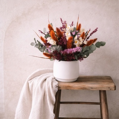 Arranjo de flores artificiais coloridas em vaso branco sobre banco de madeira com tecido branco