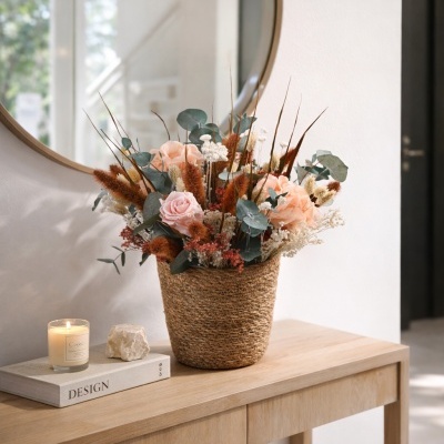 Arranjo floral com rosas cor de pêssego, flores secas e vaso de palha numa consola de madeira clara com vela e livro DESIGN.