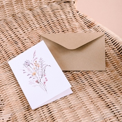 Cartão branco com desenho floral e envelope kraft sobre cadeira de vime bege
