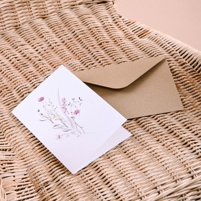 Cartão branco decorado com flores e envelope kraft sobre cadeira de vime.