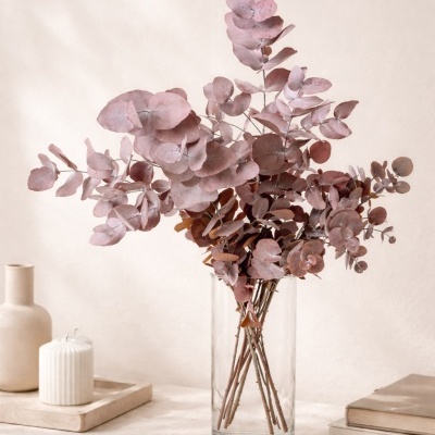 Folhagem seca rosa em vaso de vidro com vela, vaso, livros e anéis numa mesa creme.