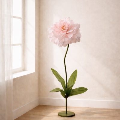 Flor decorativa rosa grande com pétalas de tecido e folhas verdes em base verde num canto iluminado.