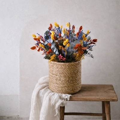Vaso em cesta de palha com flores secas coloridas sobre banco de madeira e tecido branco