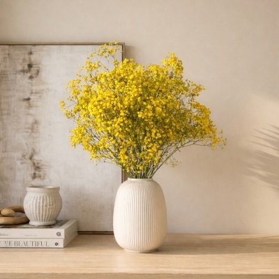 Vaso branco com ramo de flores amarelas sobre mesa de madeira com quadro e livros ao fundo