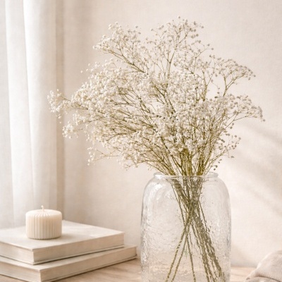 Jarro de vidro com flores brancas secas sobre mesa com livros e vela.