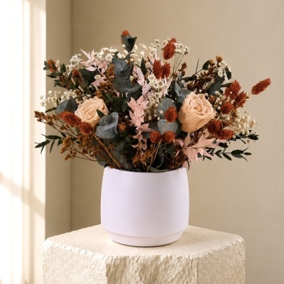Arranjo floral com rosas cor de pêssego, flores secas e folhas verdes em vaso branco sobre pedestal em tecido creme texturizado.