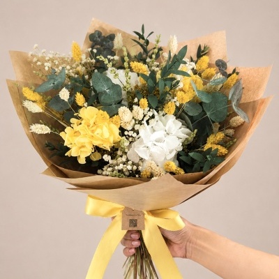 Buquê de flores secas amarelas e brancas com folhas verdes, embrulhado em papel castanho com fita amarela.