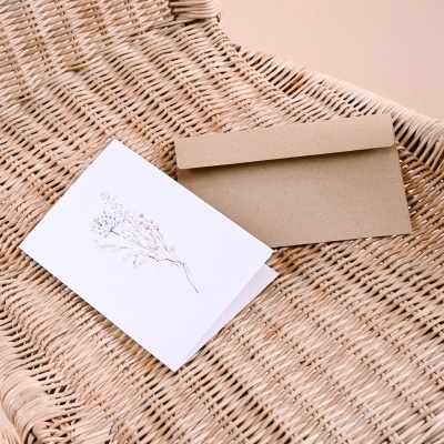 Cartão branco com flores e envelope castanho numa cadeira de vime