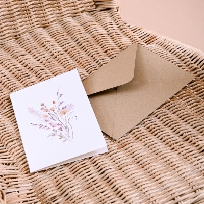Cartão branco com ilustração floral pastel e envelope castanho sobre cadeira de vime