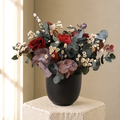 Arranjo floral em vaso preto com flores vermelhas e folhas diversas num pedestal claro.
