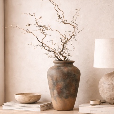 Vaso cerâmico rústico com ramos secos, taça pedra, livros e lâmpada sobre superfície de madeira clara.