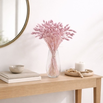 Vaso de flores secas cor-de-rosa sobre mesa de madeira com livros, tigela, vela e castiçal.