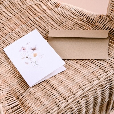 Cartão de felicitações branco com ilustração de flores e envelope castanho sobre cadeira de vime
