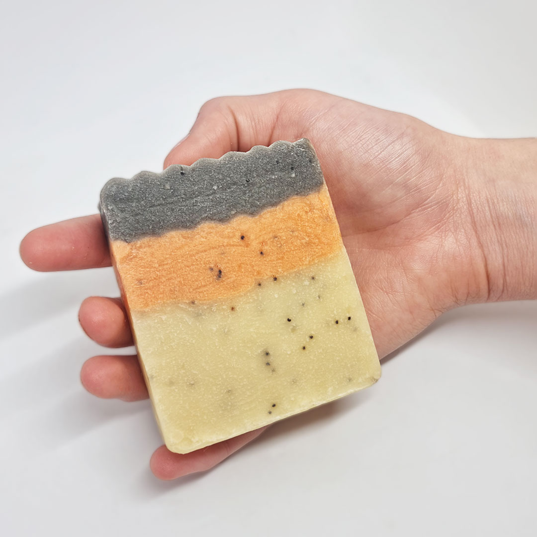 Sabonete de Âmbar e Madeiras Exóticas Sabonete artesanal com três camadas coloridas segurado por uma mão sobre fundo branco