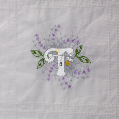 Toalha branca com bordado de letra T, flores lilás, folhas verdes e uma abelha.