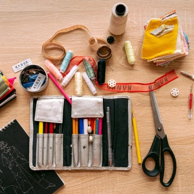 Mesa com material de costura e desenho, incluindo tesouras, fios, lápis, fita métrica vermelha e papéis com desenhos.
