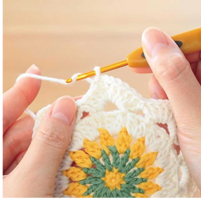 Mãos a crochetar peça floral com fio branco, amarelo e verde e agulha de crochê laranja