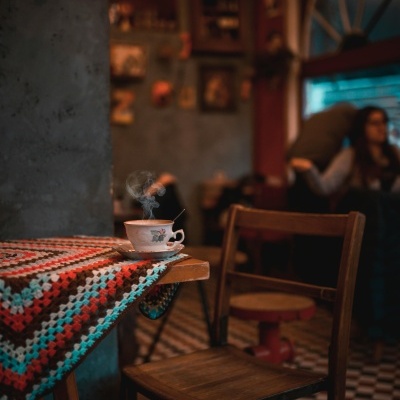 Xícara de porcelana com bebida quente numa mesa colorida de malha, cadeira de madeira, ambiente interno decorado.
