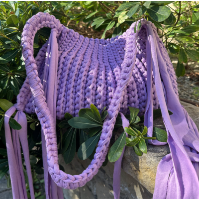 Bolsa tricotada lilás com alças trançadas e fios pendurados entre folhas verdes