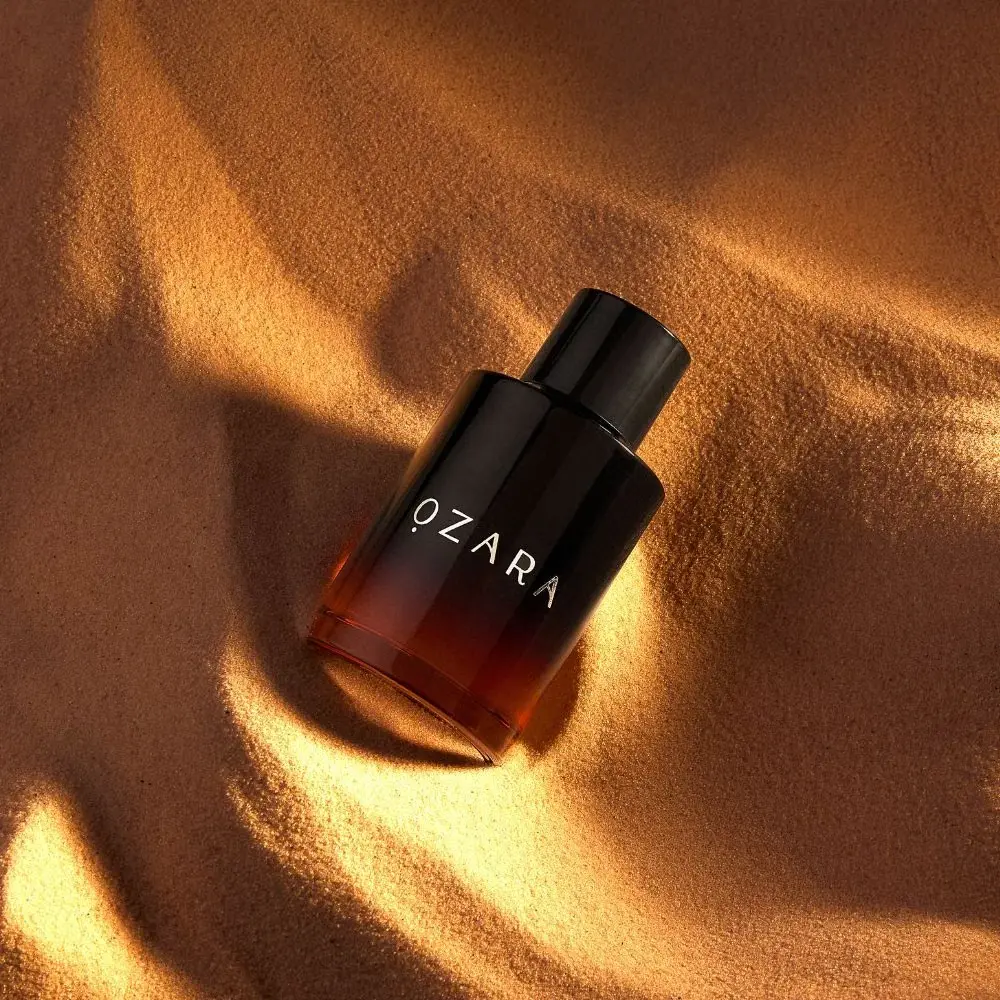 Frasco de perfume OZARA em degradê castanho para preto sobre areia dourada