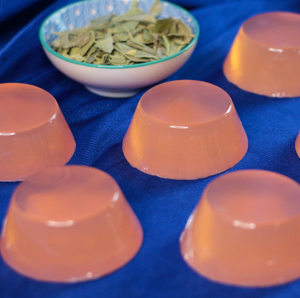 Sabonete de Boldo Seis géis translúcidos cor de pêssego sobre tecido azul com tigela de folhas verdes secas