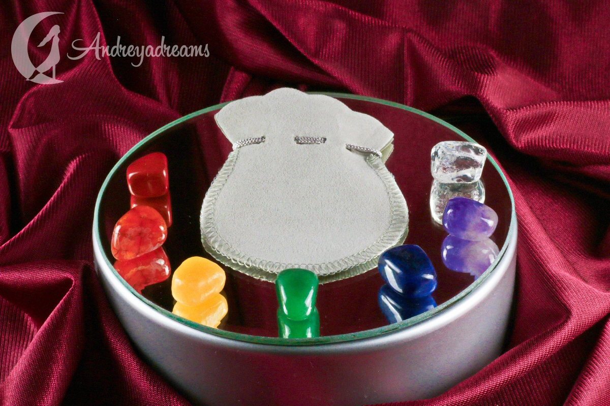 Kit Pedras dos 7 Chakras Caixa metálica redonda com tampa espelhada, pedras coloridas e saquinho cinza sobre tecido vermelho.