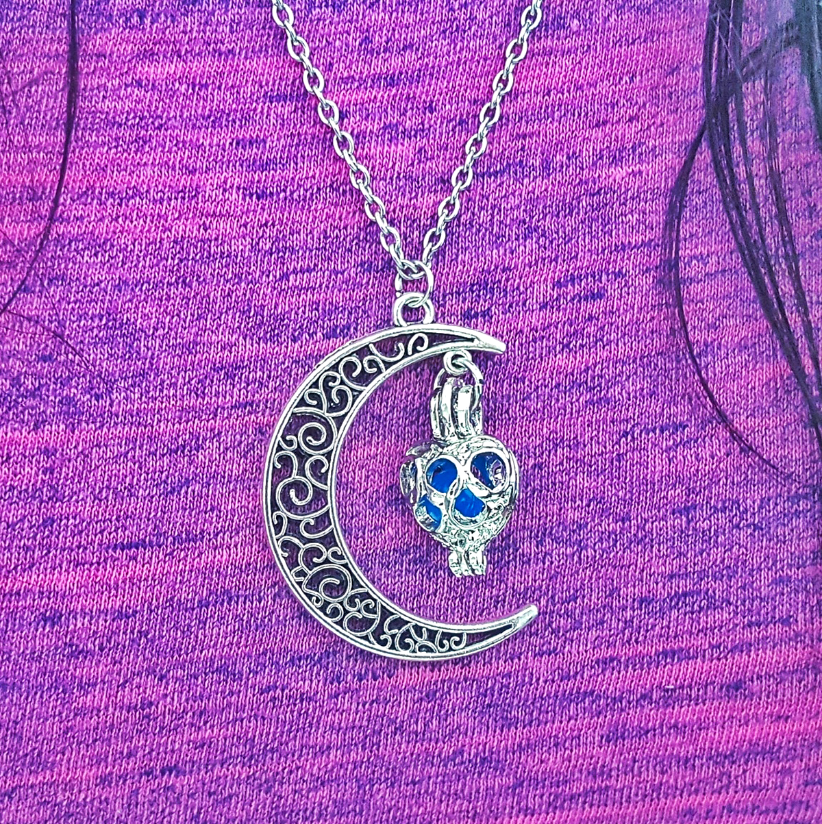 Colar Lua com Bola Que Brilha no Escuro Colar prateado com pendente de lua e crânio azul sobre roupa roxa.
