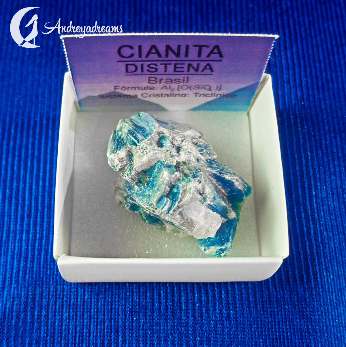 Cianite Azul Cianita azul clara em caixa branca com cartão informativo e fundo azul.