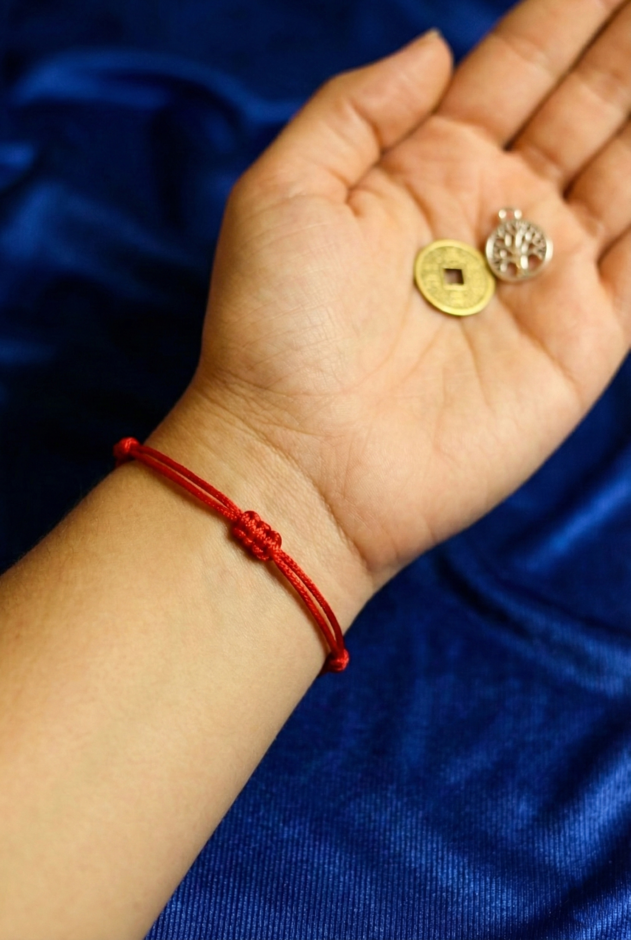 Kit Ano Novo Pulseira vermelha de cordão com nó central na mão segurando moeda dourada e pendente prateado sobre tecido azul.