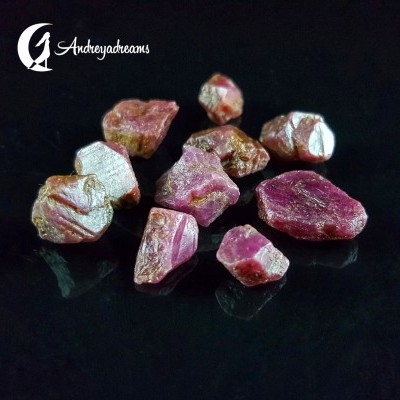 Pedras preciosas brutas vermelhas e rosas sobre superfície preta, com marca AndreyaDreams