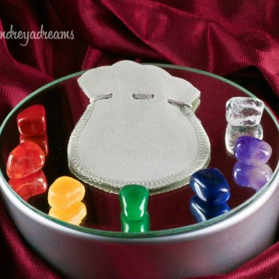 Caixa metálica redonda com tampa espelhada, pedras coloridas e saquinho cinza sobre tecido vermelho.