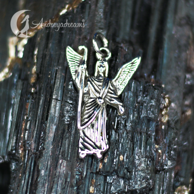 Pendente em metal prateado em forma de anjo com asas, sobre superfície escura texturizada