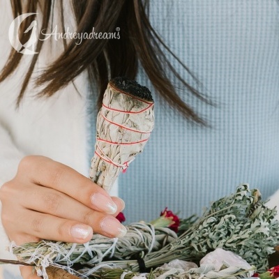 Mão a segurar feixe de sálvia branca amarrado com fio vermelho com ervas secas no fundo