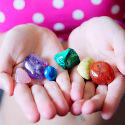 Mãos segurando seis pedras polidas coloridas com vestido rosa de bolinhas brancas ao fundo