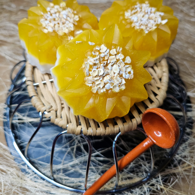 Sabonetes amarelos em forma de flor com aveia no centro sobre suporte redondo e colher pequena de madeira