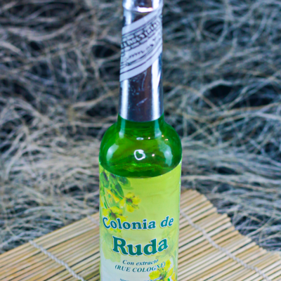 Garrafa verde com rótulo amarelo e verde de Colonia de Ruda sobre esteira de madeira clara.