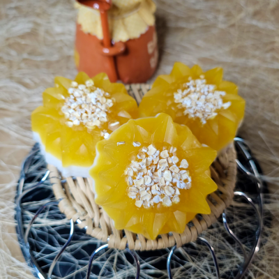 Três sabonetes amarelos em forma de girassol sobre base de rattan e metal, boneca de barro ao fundo