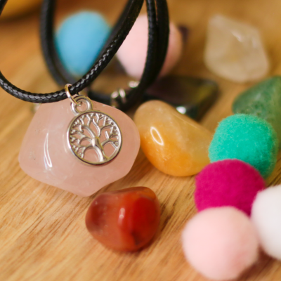 Colar com pingente de pedra rosa e medalha prateada com desenho de árvore sobre superfície de madeira com pedras coloridas e pompons de lã.