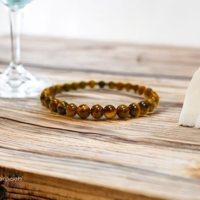 Pulseira de contas de pedra castanho-verde sobre madeira clara com taça de vidro ao fundo