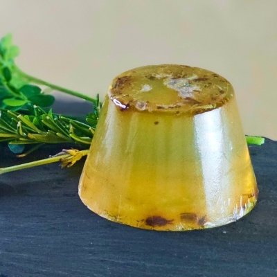 Sabonete translúcido amarelo com ervas sobre superfície escura e raminhos verdes