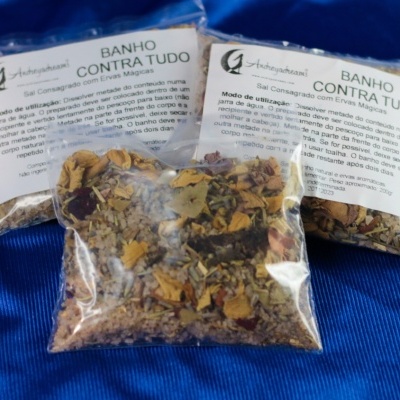 Pacotes transparentes com mistura de ervas secas rotulados com texto BANHO CONTRA TUDO sobre tecido azul